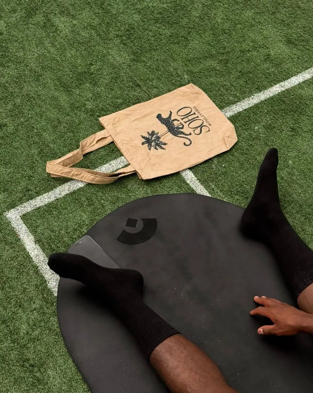 Soho Boulevard tote bag beside a member stretching on an exercise mat on the artificial turf